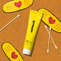 Yellow tube of emimerk Centella Salve on an orange background with cartoon bandages and cotton swabs.
